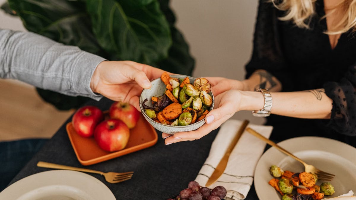 Two hands sharing a bowl of colorful roast vegetables on a table with plates, fruits, and cutlery.
